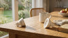 A sunlit wooden table features two LED bulbs, a steaming ceramic mug, a bowl of fruit, and a book, creating a cozy, inviting atmosphere.