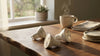 Three LED bulbs lie on a wooden table next to a steaming coffee cup and stacked plates, with sunlight streaming through a window and a potted plant nearby.
