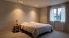 A cozy bedroom with soft lighting featuring a neatly made double bed, a wooden nightstand with a lamp, and a window with sheer curtains letting in daylight.