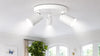 A modern white ceiling light fixture with three bright spotlights illuminating a cozy room, featuring framed butterfly art and large windows with greenery outside.